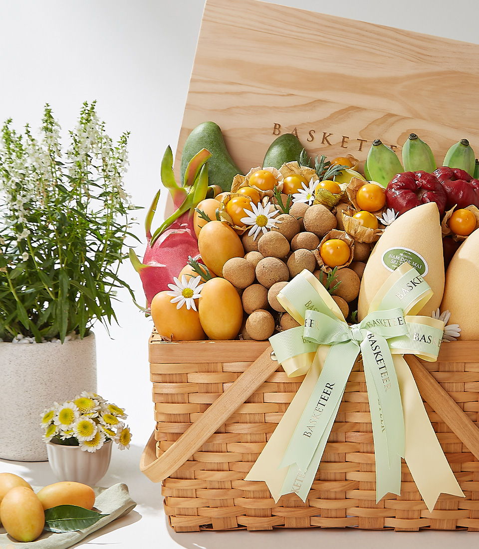 A wicker basket filled with premium Mayongchids from Nakhon Nayok, Nam Dok Mai See Thong mangoes, Keo Seowei green mangoes, red skin dragon fruit, Ducasse bananas, rose apples, longans, and cape gooseberries, elegantly arranged with a green ribbon and wooden lid