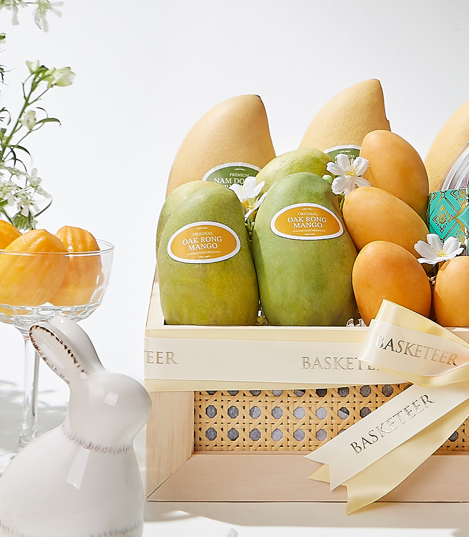 A rectangular wooden gift basket with cane details, filled with premium Mayongchids from Nakhon Nayok, Nam Dok Mai See Thong mangoes, Ok-Rong mango, and Fairy Dust sticky rice, elegantly decorated with a cream ribbon and white flowers