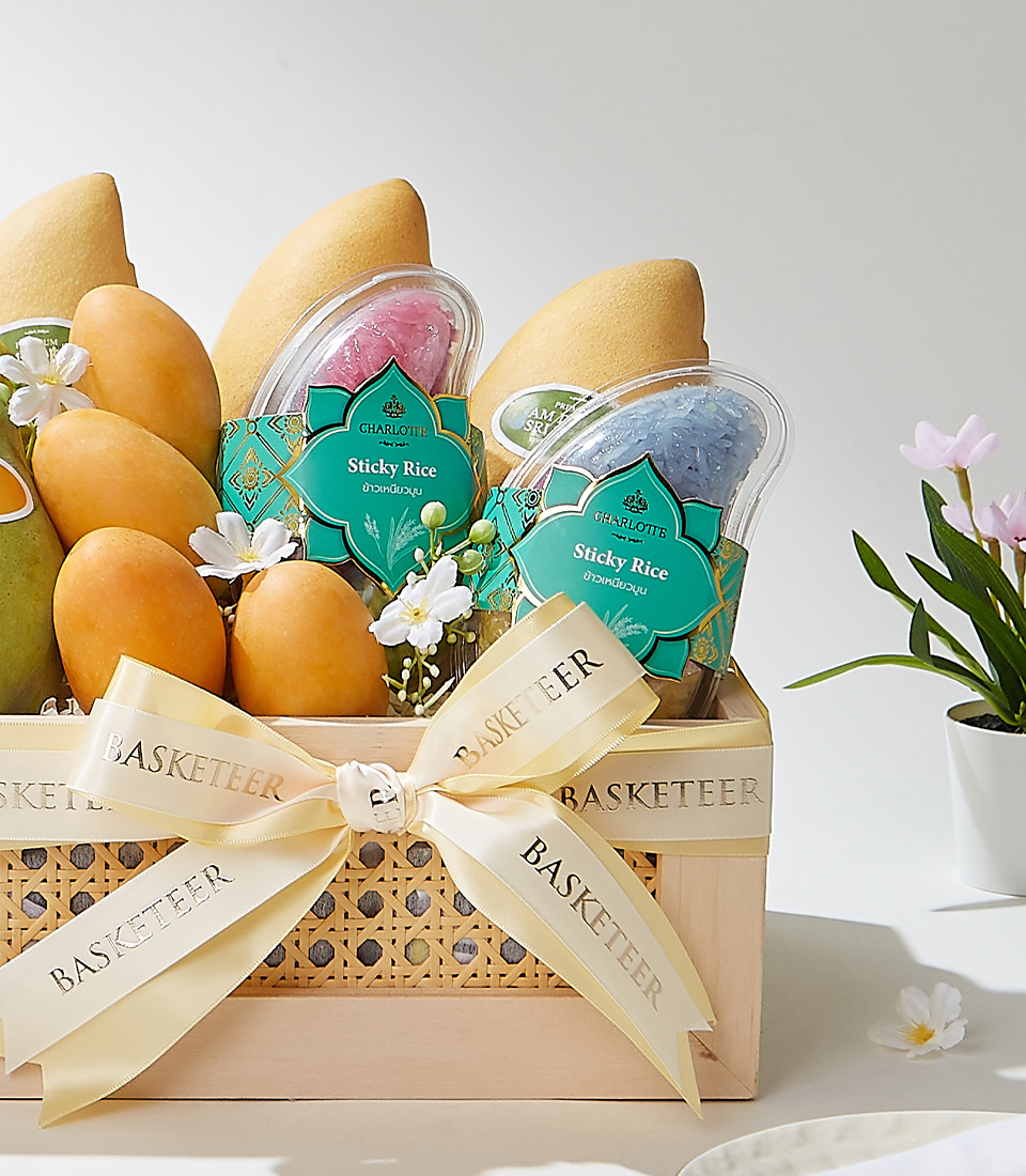 A rectangular wooden gift basket with cane details, filled with premium Mayongchids from Nakhon Nayok, Nam Dok Mai See Thong mangoes, Ok-Rong mango, and Fairy Dust sticky rice, elegantly decorated with a cream ribbon and white flowers