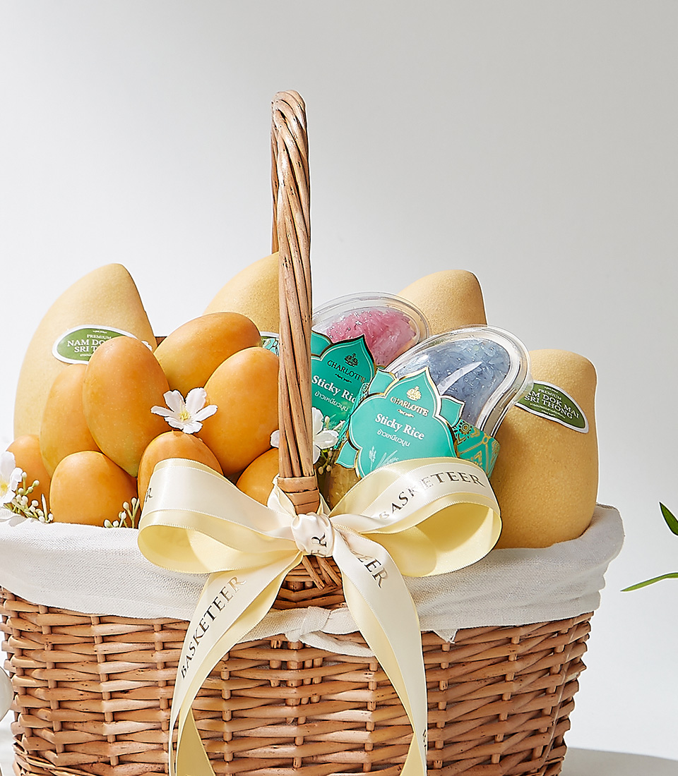 A Classic Grace wicker basket filled with premium Mayongchids from Nakhon Nayok, Nam Dok Mai See Thong mangoes, and Fairy Dust sticky rice, elegantly decorated with a cream ribbon and white flowers