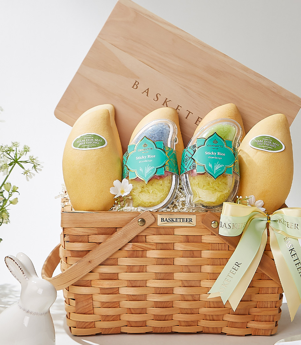 A wooden picnic basket set with handles, filled with Nam Dok Mai See Thong mangoes from Chachoengsao and Fairy Dust sticky rice, elegantly arranged with a green ribbon and fresh flowers