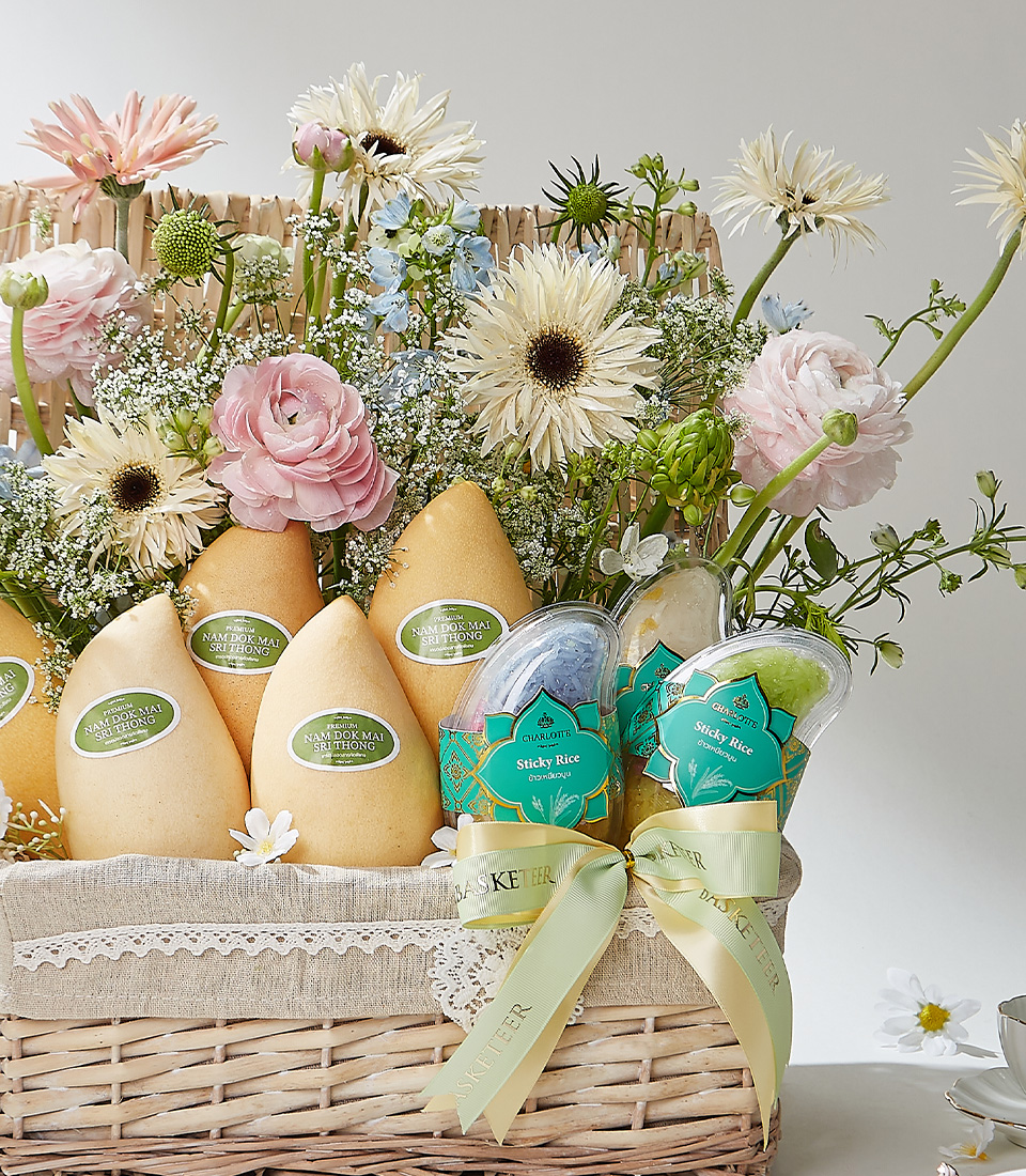 A Rustic Lace storage basket filled with Nam Dok Mai See Thong mangoes from Chachoengsao, Fairy Dust sticky rice, and Snow Pearl sticky rice, elegantly arranged with fresh flowers and a green ribbon