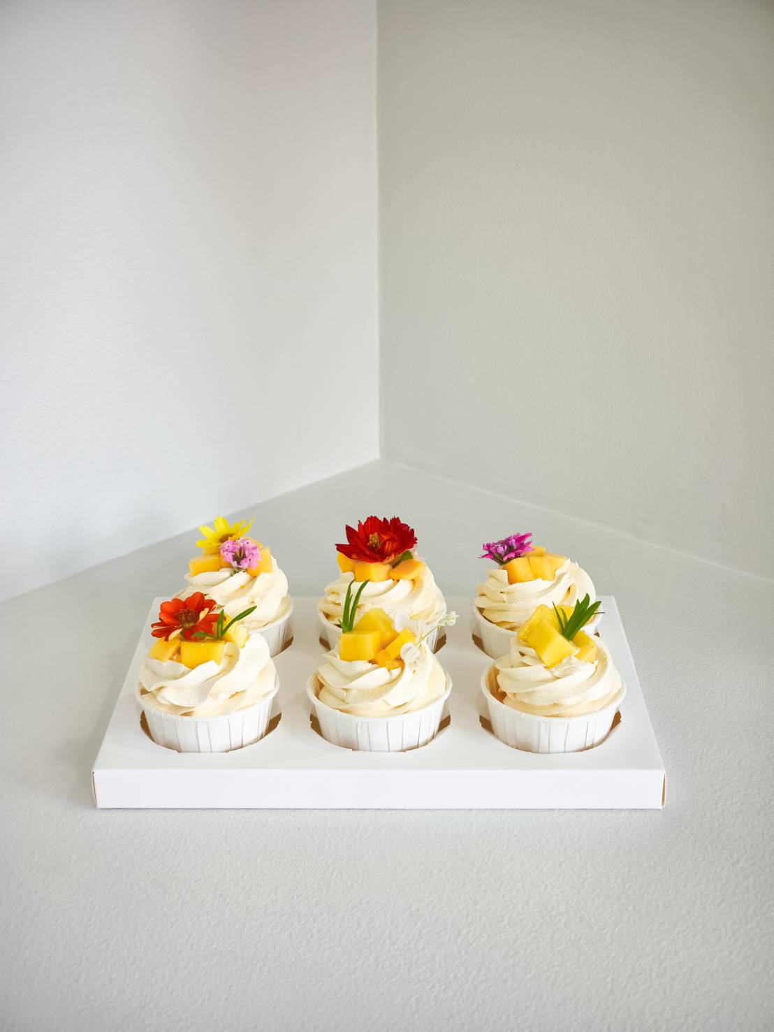 Set of six vanilla cupcakes topped with fresh mango cubes and colourful edible flowers, arranged in a white box.