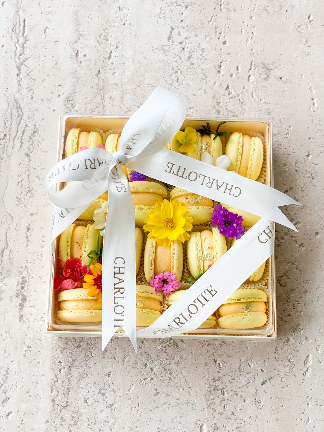 An elegant Charlotte Bakery box of yellow and light green macarons with fresh flowers and ribbon, a refined treat in Bangkok.