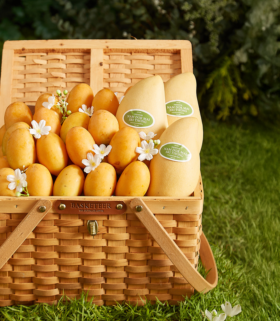 Close-up of a beautifully woven picnic basket filled with premium Mayongchids (Toon Klaow Variety Nakhon Nayok) and Nam Dok Mai See Thong Mangoes (Chachoengsao), adorned with delicate white flowers.