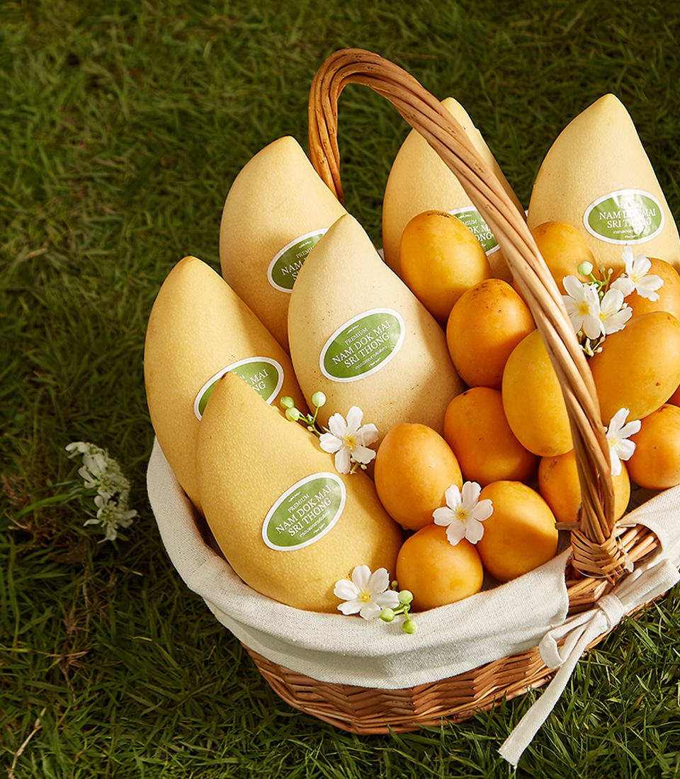 Close-up of a beautifully arranged wicker basket filled with Premium Mayongchids (Toon Klaow Variety Nakhon Nayok) and Nam Dok Mai See Thong Bang Khla Mangoes, decorated with delicate white flowers, set on a lush green grass background.