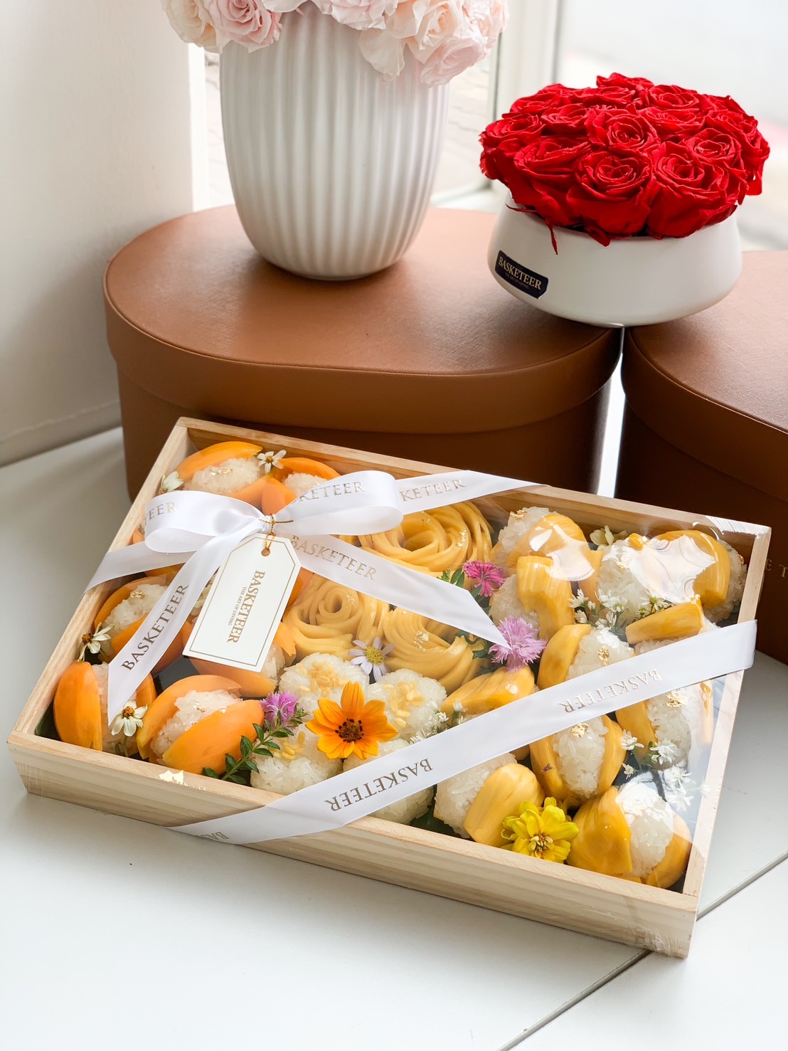 A luxurious gift box filled with premium Thai desserts, including golden mango rosettes, Mayongchid (Marian Plum) sticky rice, and jackfruit sticky rice, elegantly arranged with edible flowers. The box is wrapped with a white ribbon and gold-lettered tag from Basketeer, making it a perfect gift for special occasions.
