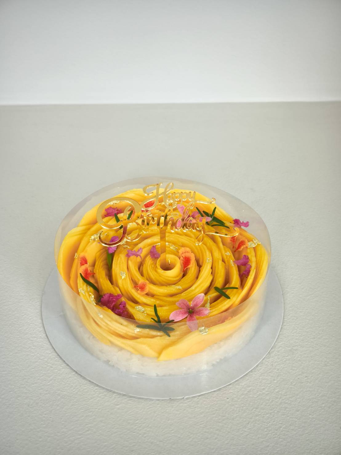Mango sticky rice birthday cake topped with fresh mango slices, edible flowers, gold leaf, and Happy Birthday topper