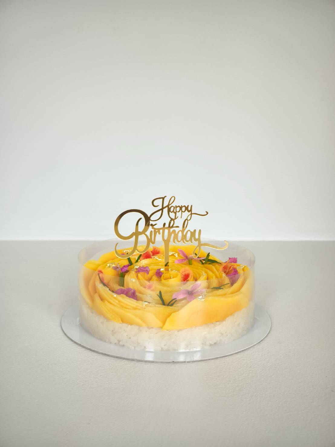 Mango sticky rice birthday cake with golden Happy Birthday topper, fresh mango rosettes, edible flowers, and glutinous rice base
