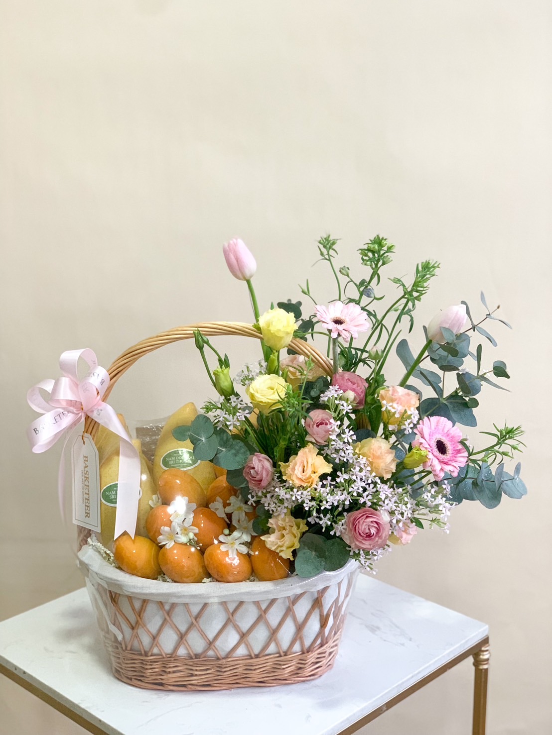 Pastel fruit and flower basket with fresh mangoes and delicate pink, peach, and white floral arrangement in a wicker basket.