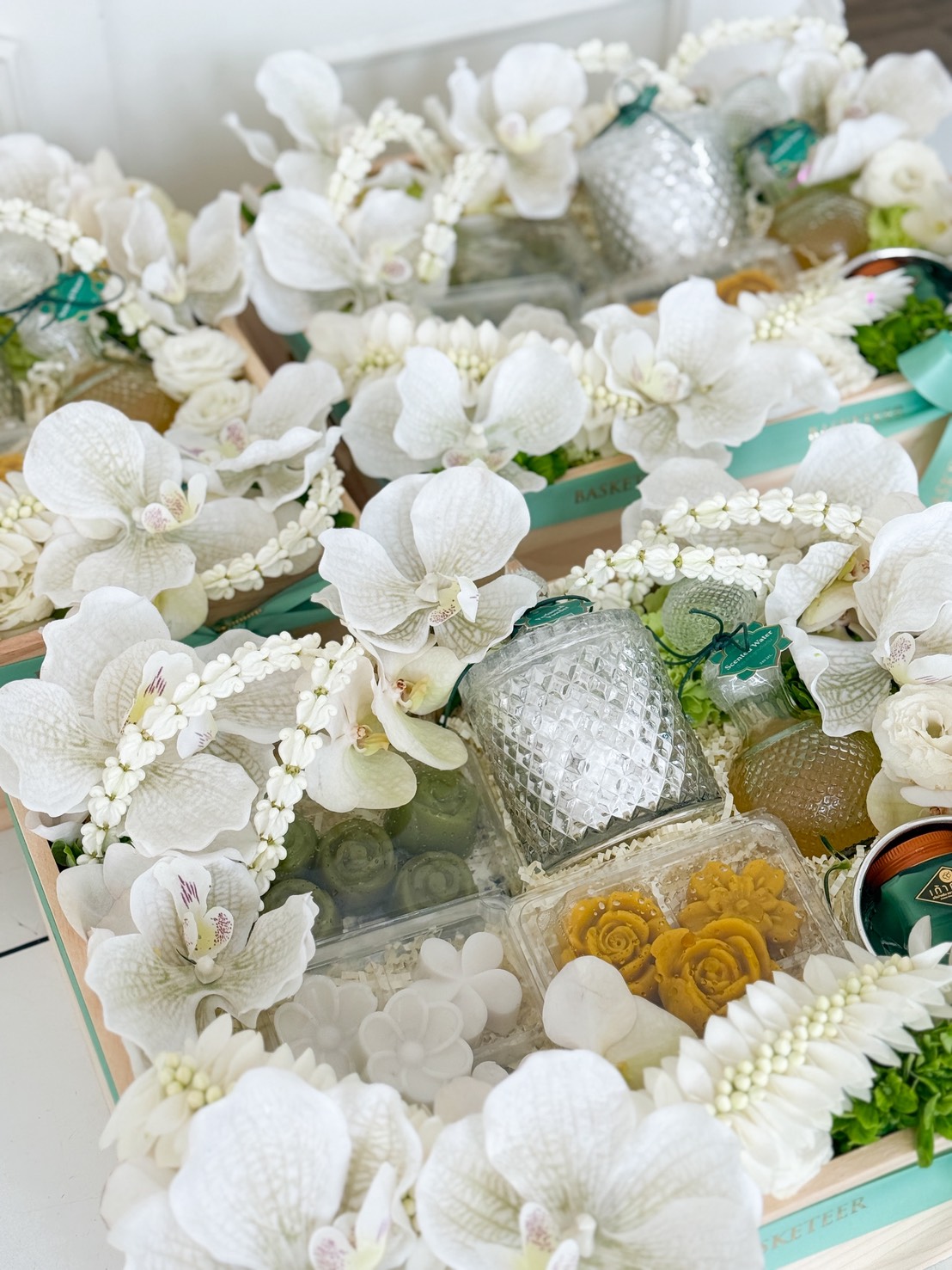 Gift hampers decorated with white orchids and flowers, filled with glass jars, packaged sweets, and green and yellow treats—our Thai Sweets & Good Fortune Songkran Set is arranged in a decorative and elegant presentation.