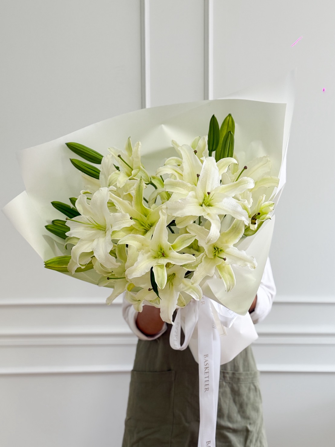 A person holding the Pure Elegance White Lily Bouquet, featuring a large arrangement of white lilies wrapped in white paper with a white ribbon, stands against a plain white wall.