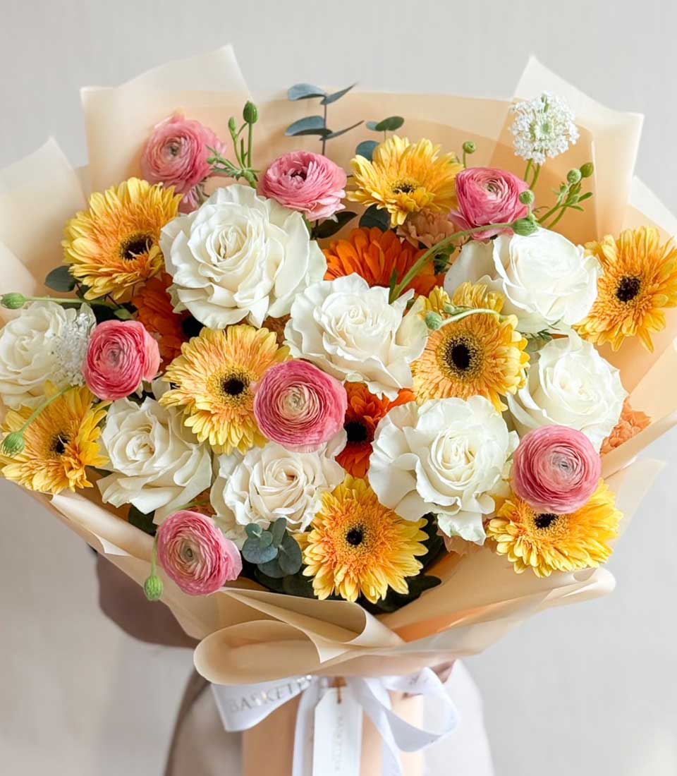 A bright and cheerful bouquet with white roses, yellow and orange gerberas, and pink ranunculus — wrapped in soft cream paper with Basketeer ribbons.