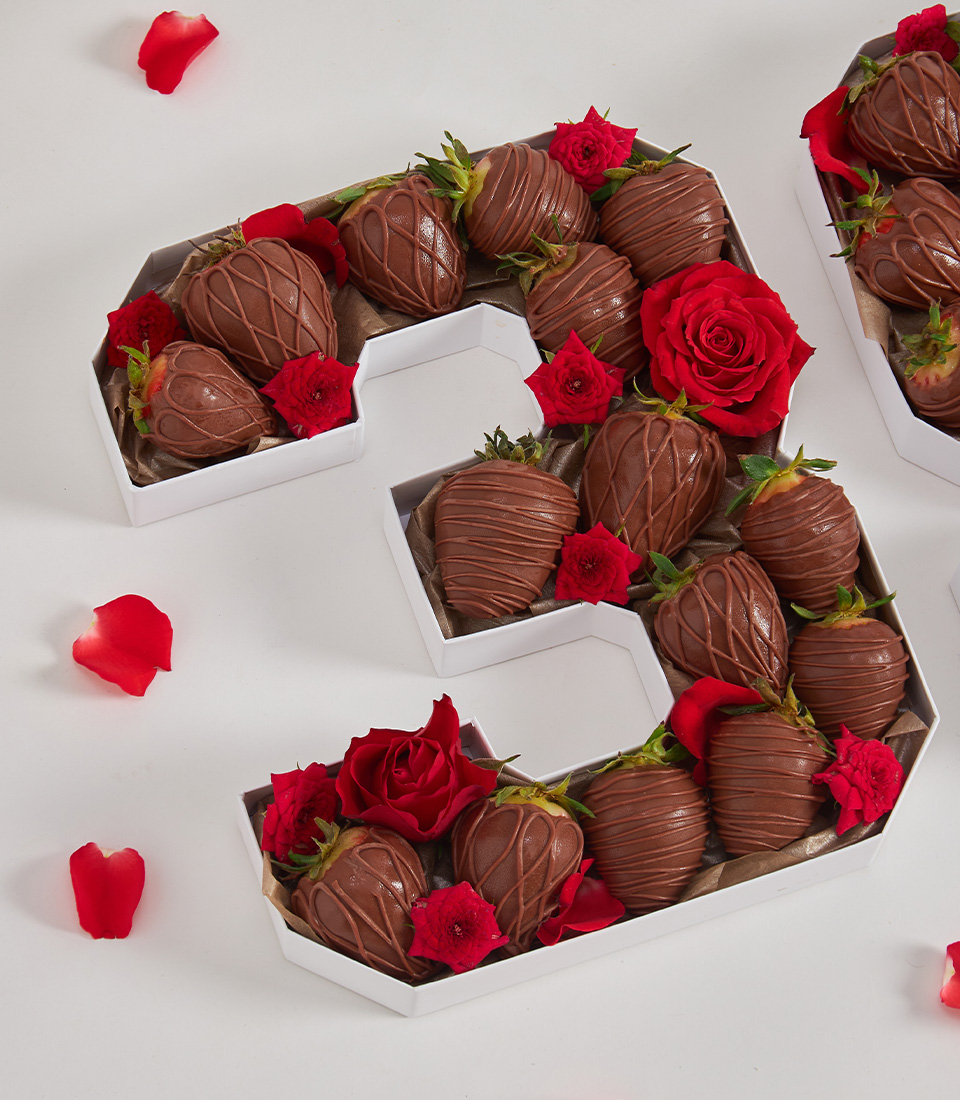 Close-up of number 3 box with chocolate strawberries and red roses on a white background with scattered rose petals