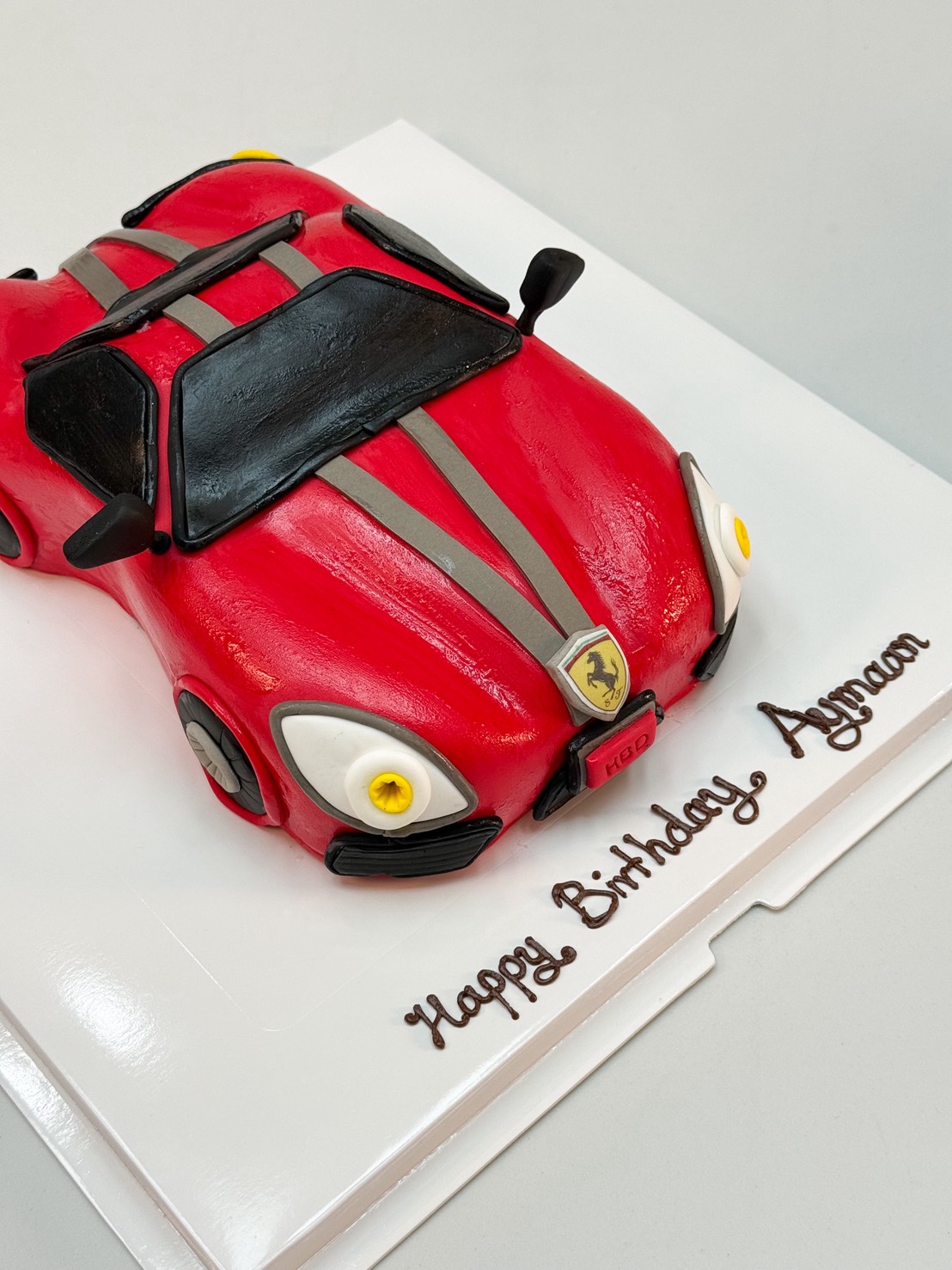 3D red sports car birthday cake with grey racing stripes, handcrafted details, on a white cake board with chocolate “Happy Birthday” message.