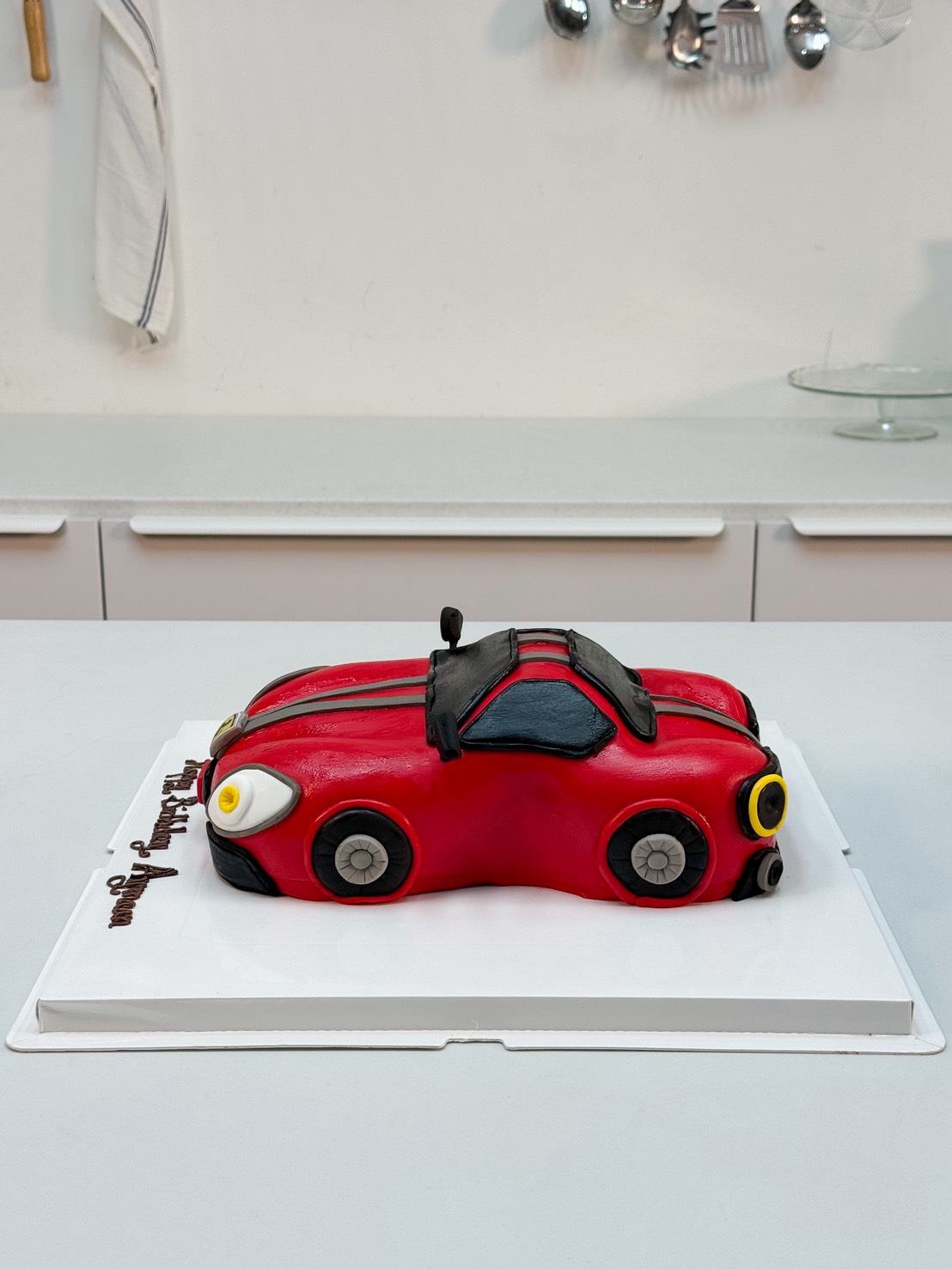 A Red Sports Car Cake (4 lb), shaped and decorated with black, yellow, and white accents, sits on a white tray in a modern kitchen.
