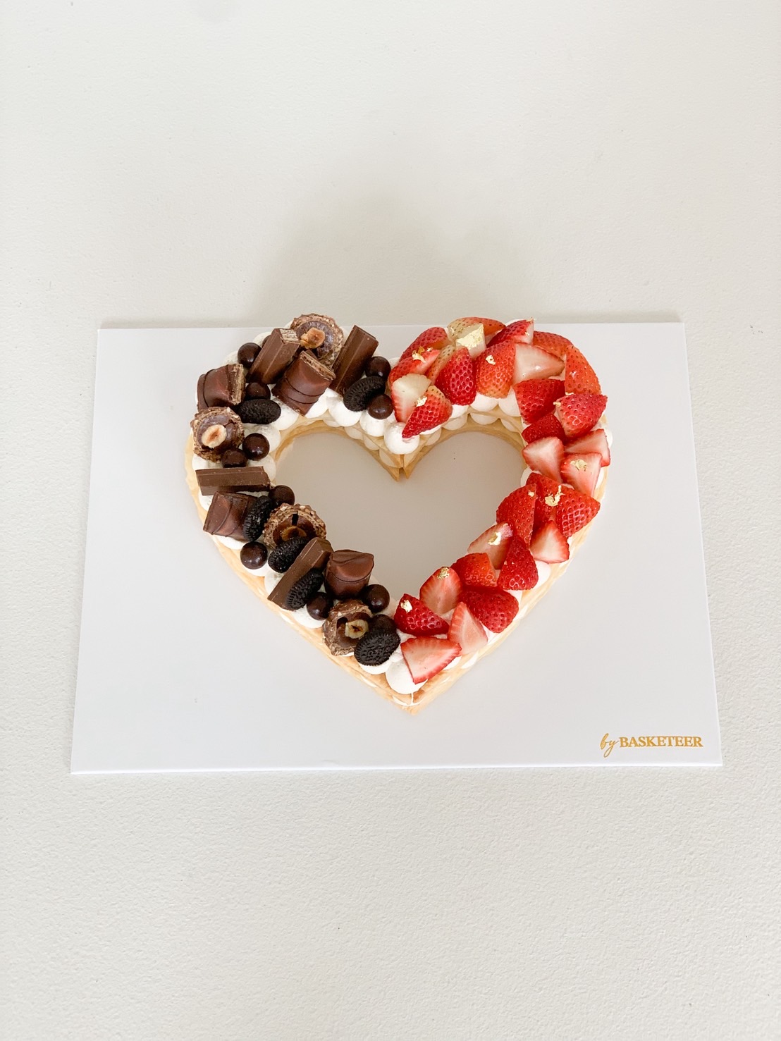 Heart-shaped cookie cake topped with Belgian chocolates and fresh strawberries on white board.