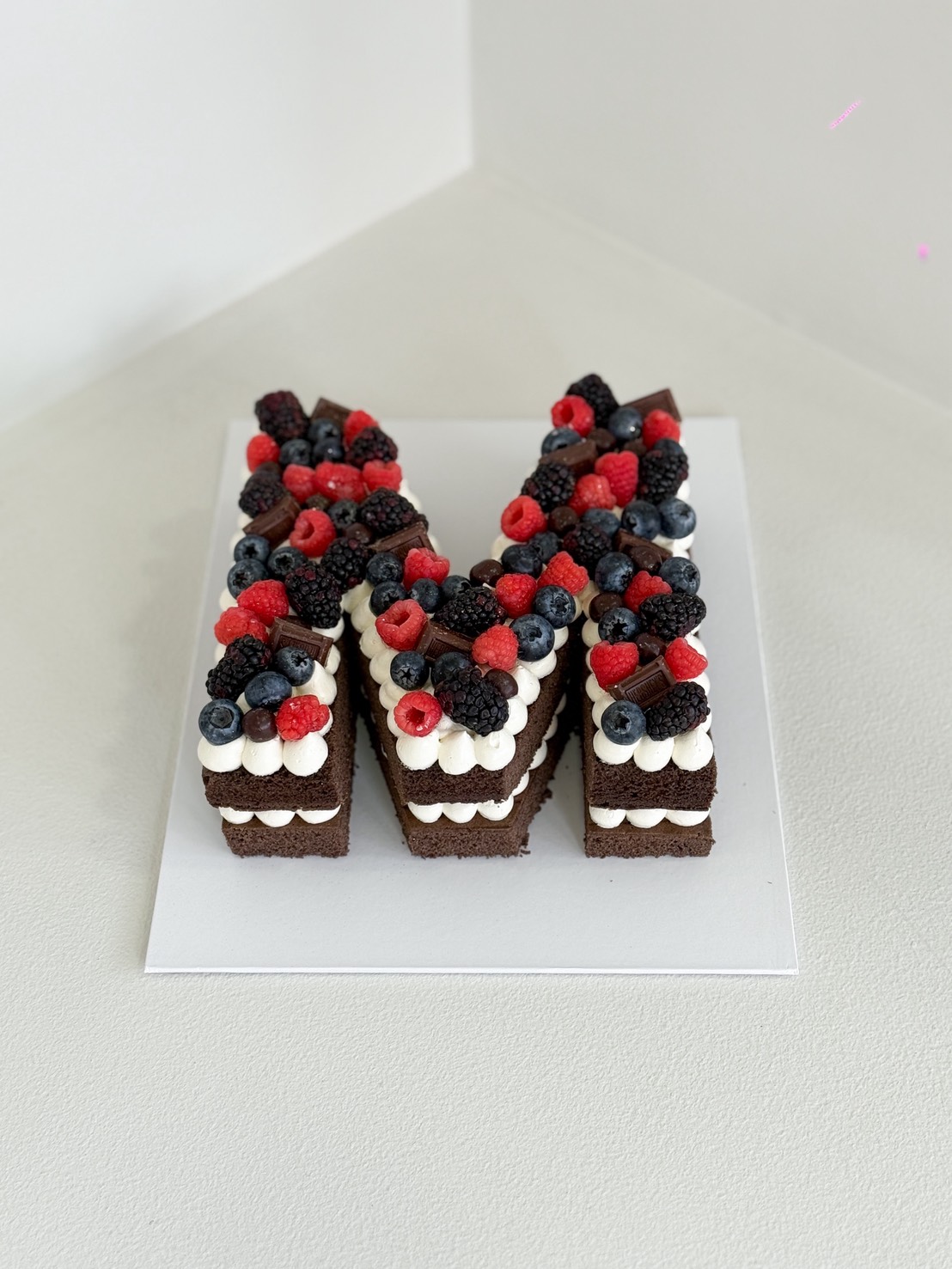 Chocolate letter M cake topped with fresh raspberries, blueberries, blackberries, and chocolate pieces, layered with cream frosting