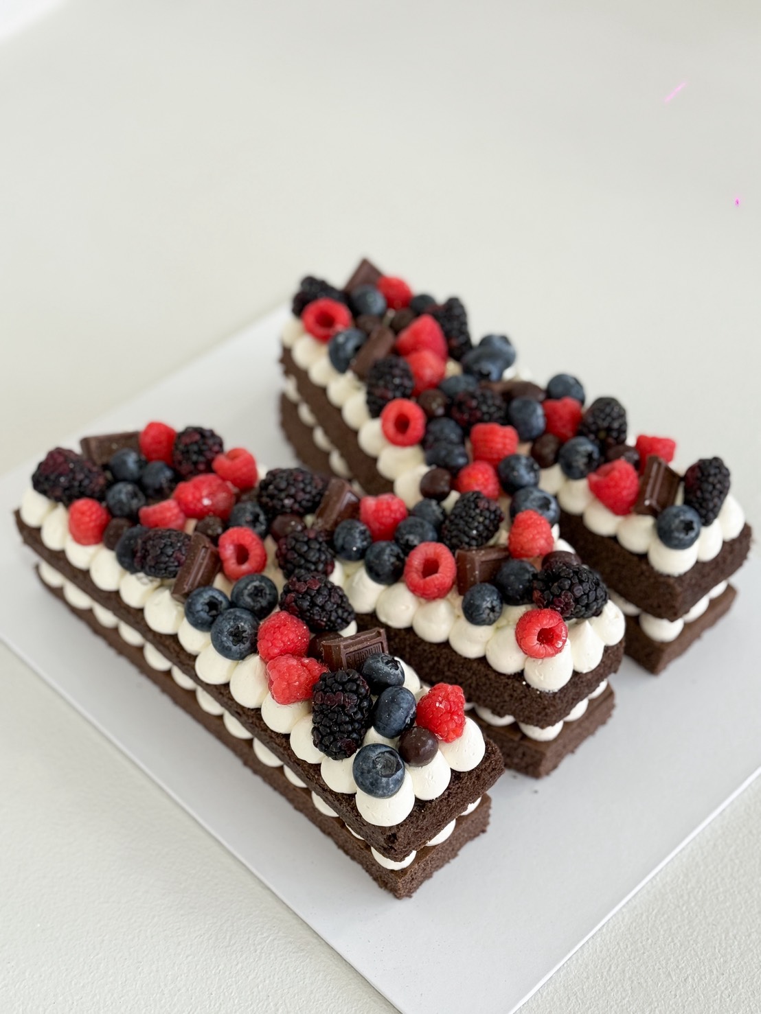 Chocolate letter H cake layered with cream frosting, decorated with raspberries, blueberries, blackberries, and chocolate chunks