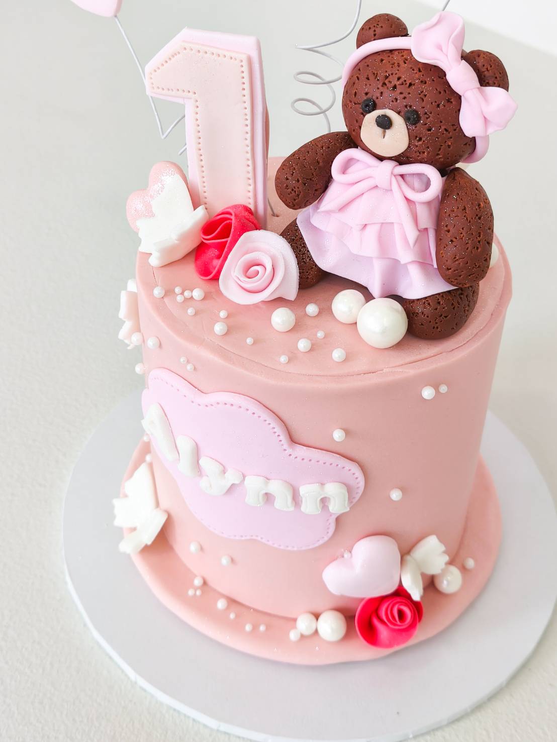 A pink birthday cake adorned with a fondant teddy bear, roses, and “Alynn Charlotte” plaque, crafted in Bangkok by Charlotte Bakery.