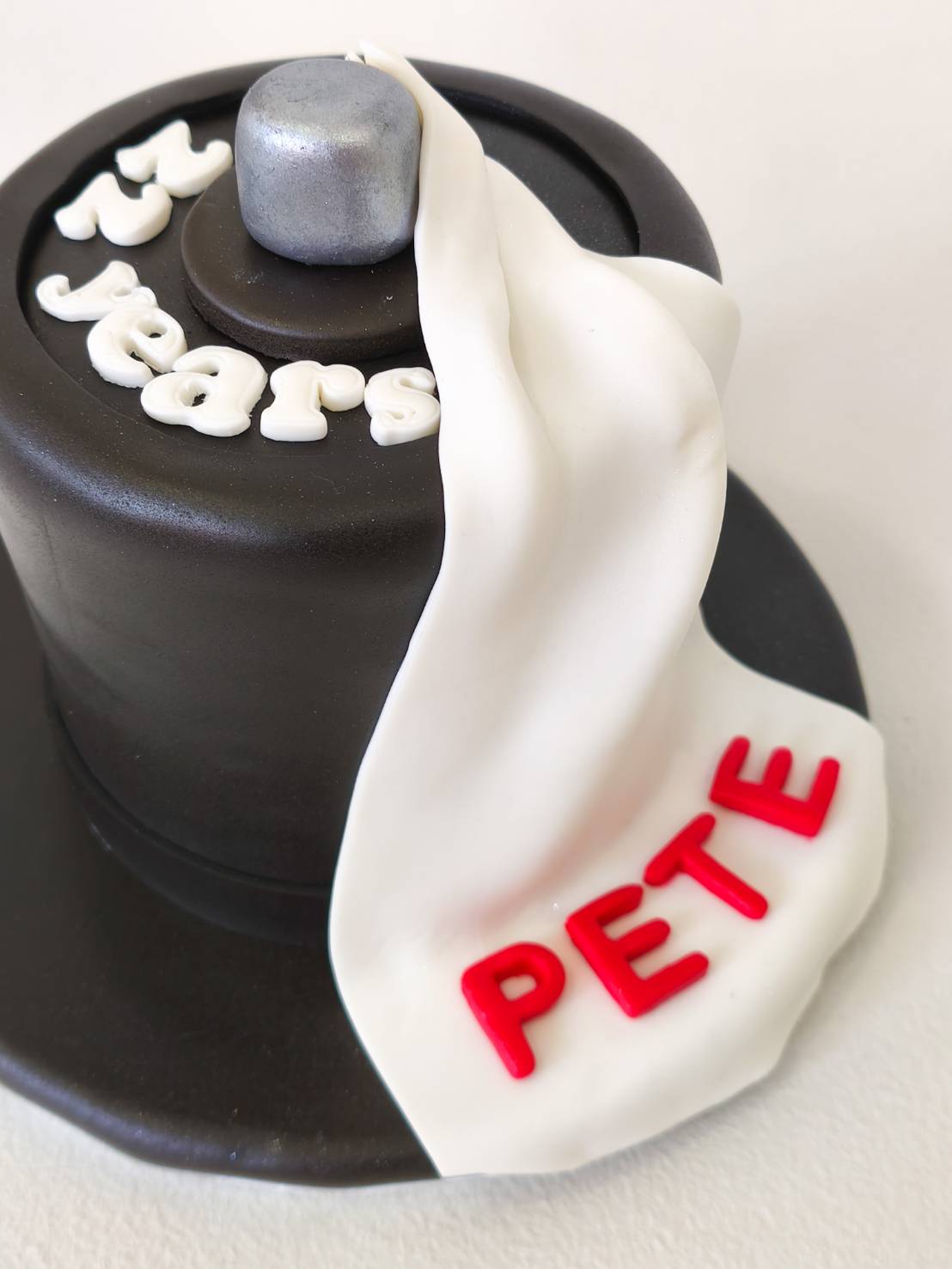 Black fondant cake shaped like a top hat with “22 years” lettering and a white fondant scarf reading “PETE” in red.