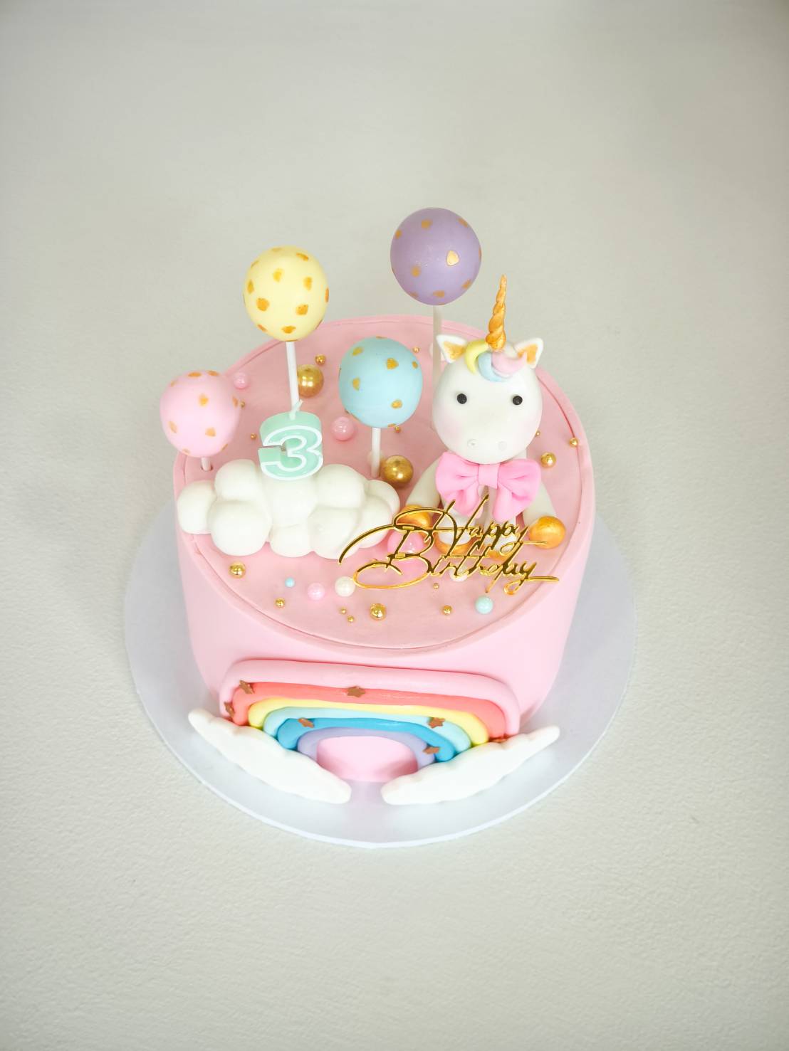 Pink unicorn birthday cake with rainbow decoration, fondant balloons, clouds, and a gold “Happy Birthday” topper.