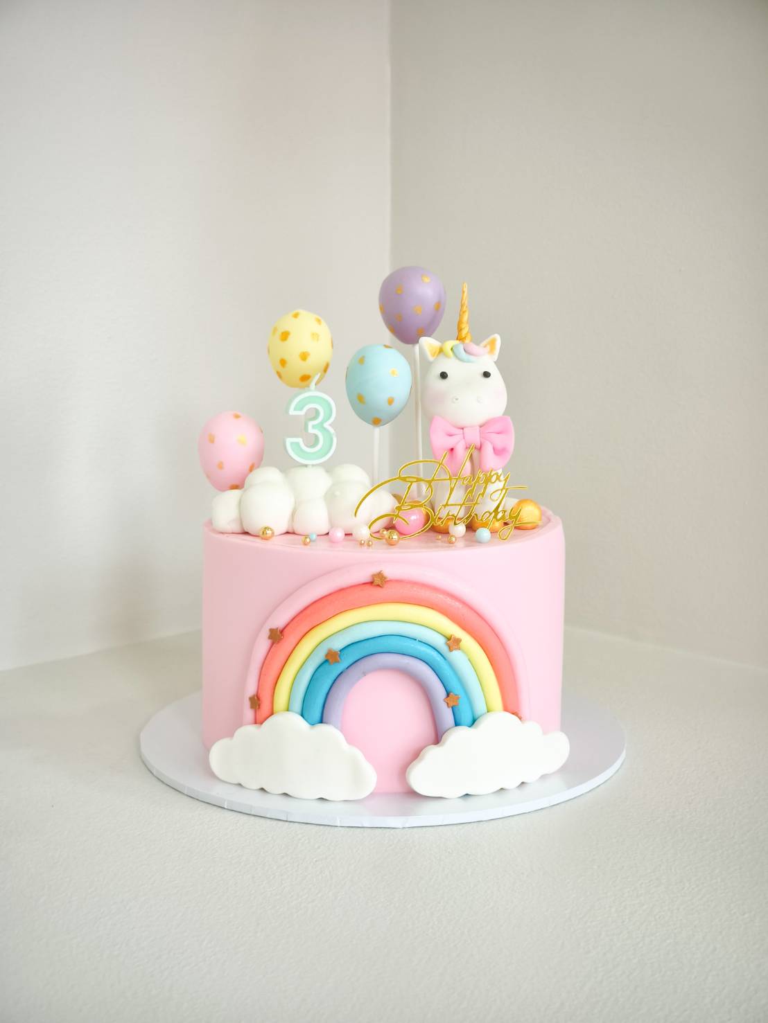 Pink unicorn birthday cake with rainbow and cloud fondant design, pastel balloons, and gold “Happy Birthday” topper.