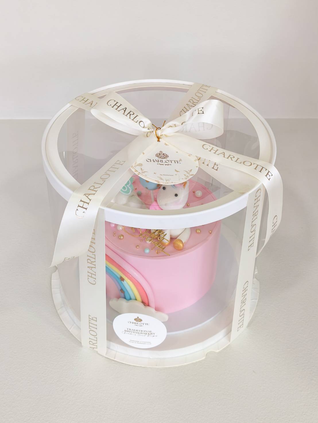 A charming pink Charlotte cake with a unicorn topper, gold details, and rainbow, presented in a clear round box with white ribbon.