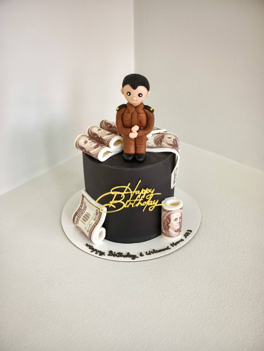 Black birthday cake decorated with edible money sheets and a fondant figure of a Thai officer in brown uniform, with gold “Happy Birthday” topper.