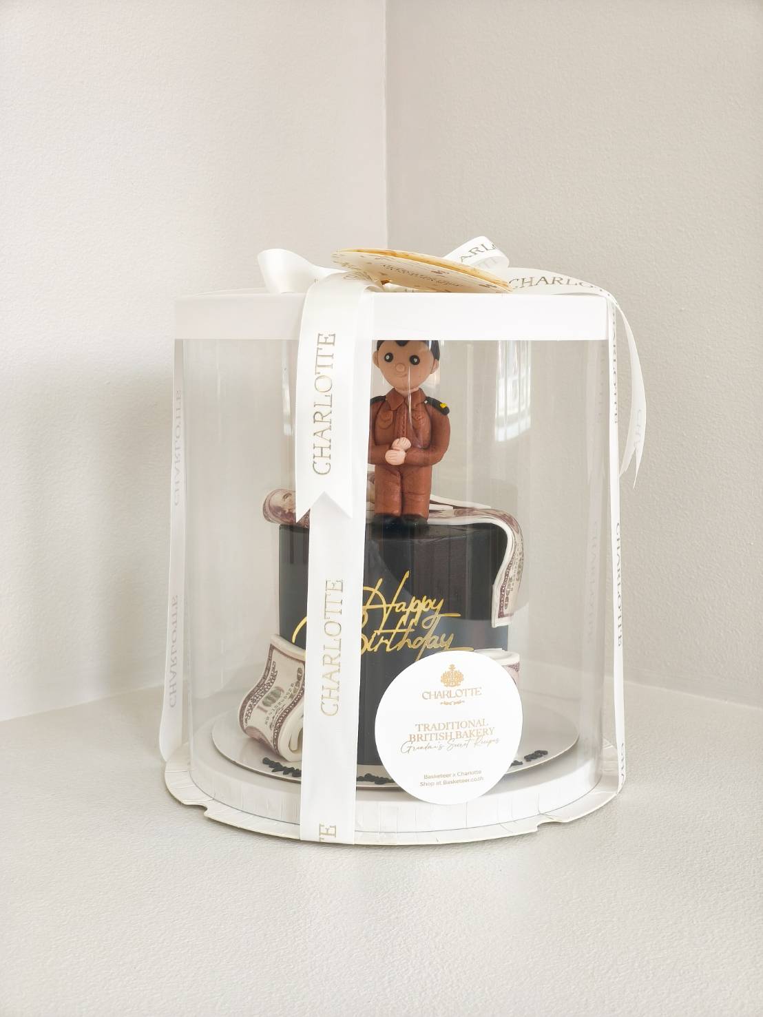 Black birthday cake with fondant Thai officer figure and money roll design, presented in a clear luxury gift box with white ribbons.