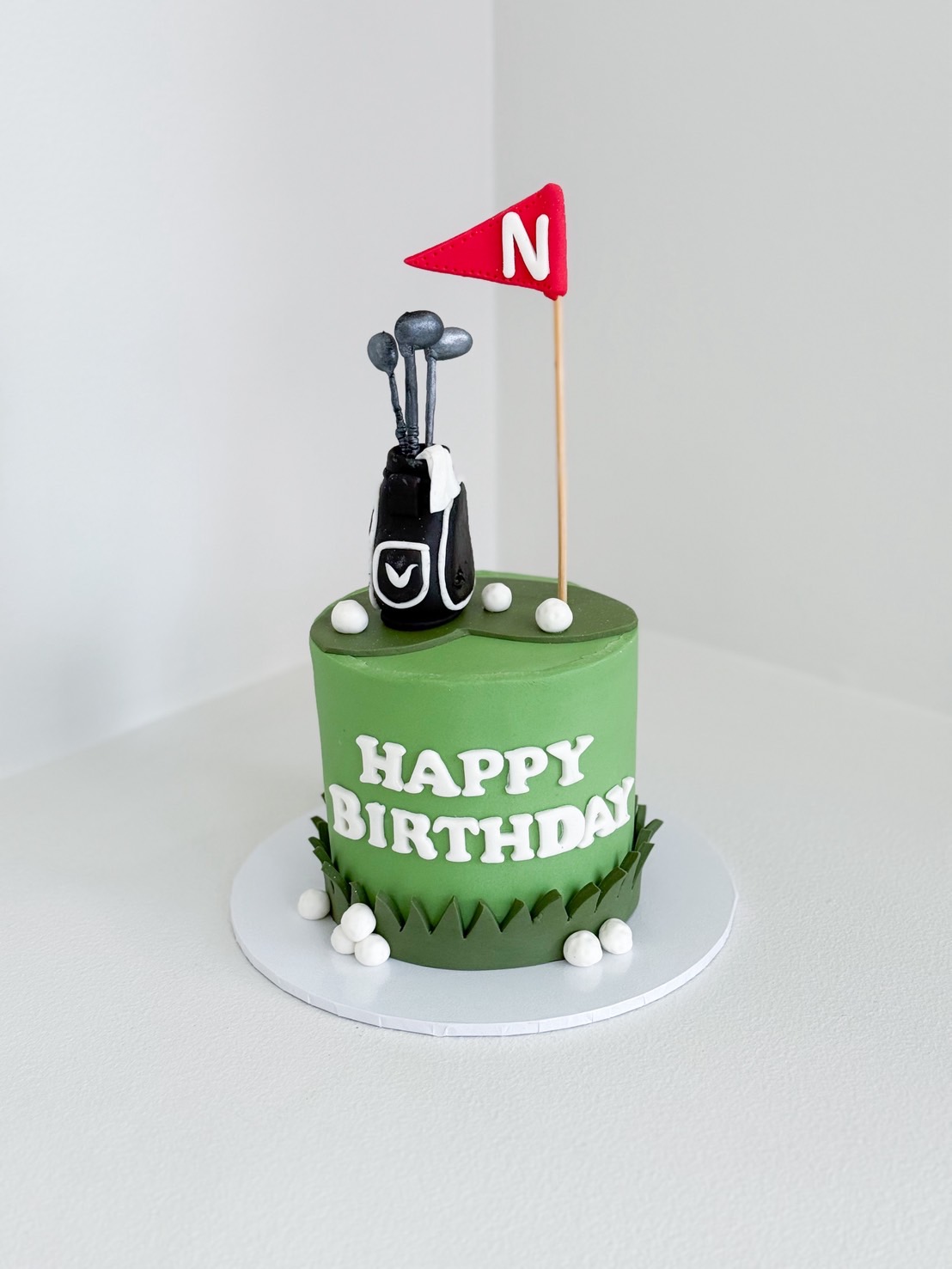 Green golf-themed birthday cake with fondant golf bag, clubs, white golf balls, and red flag topper with letter “N”.