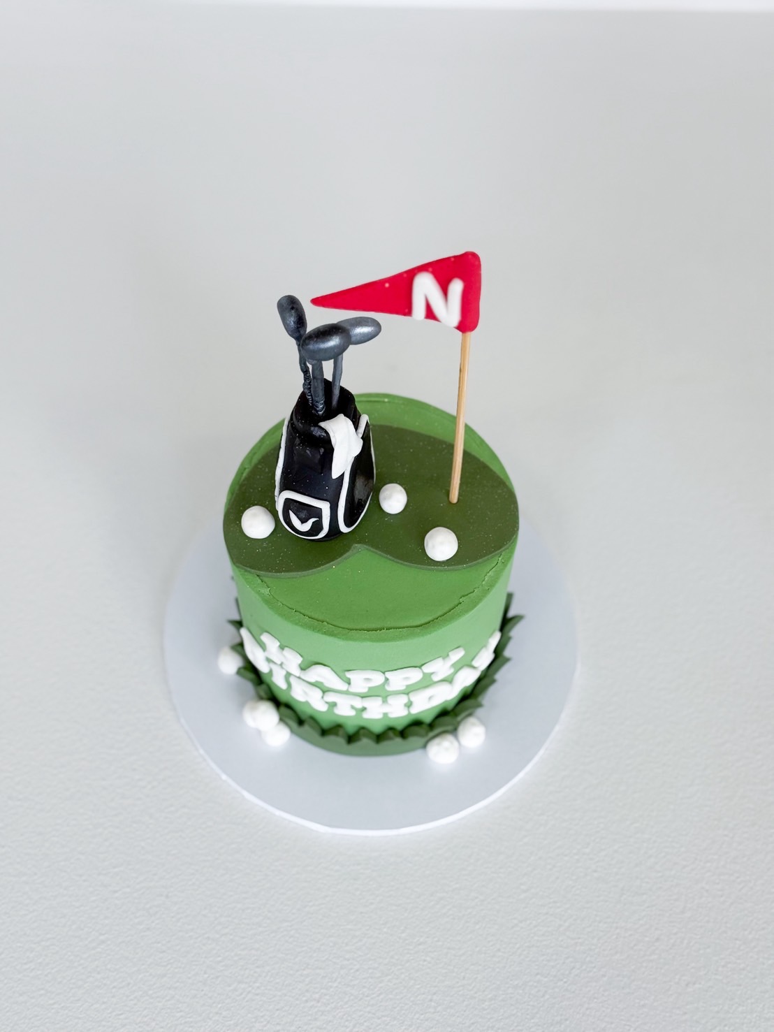 Top view of green golf-themed birthday cake with fondant golf bag, clubs, red flag, and white edible golf balls on a silver cake board.