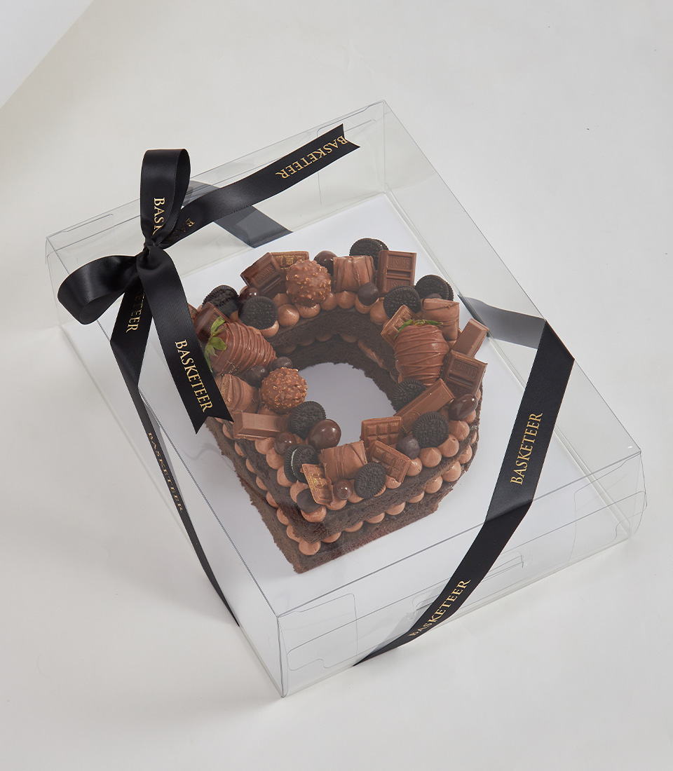Chocolate sponge letter cake topped with assorted chocolates, mini Oreos, and chocolate-dipped strawberries in a clear gift box