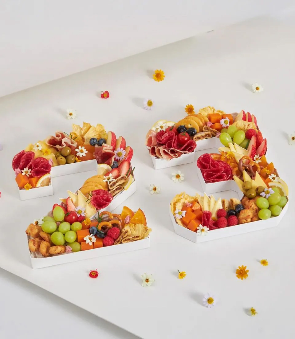 Number 23 charcuterie letter cake topped with fruits, cheeses, salami, ham, olives, pretzels, and edible flowers on a white background