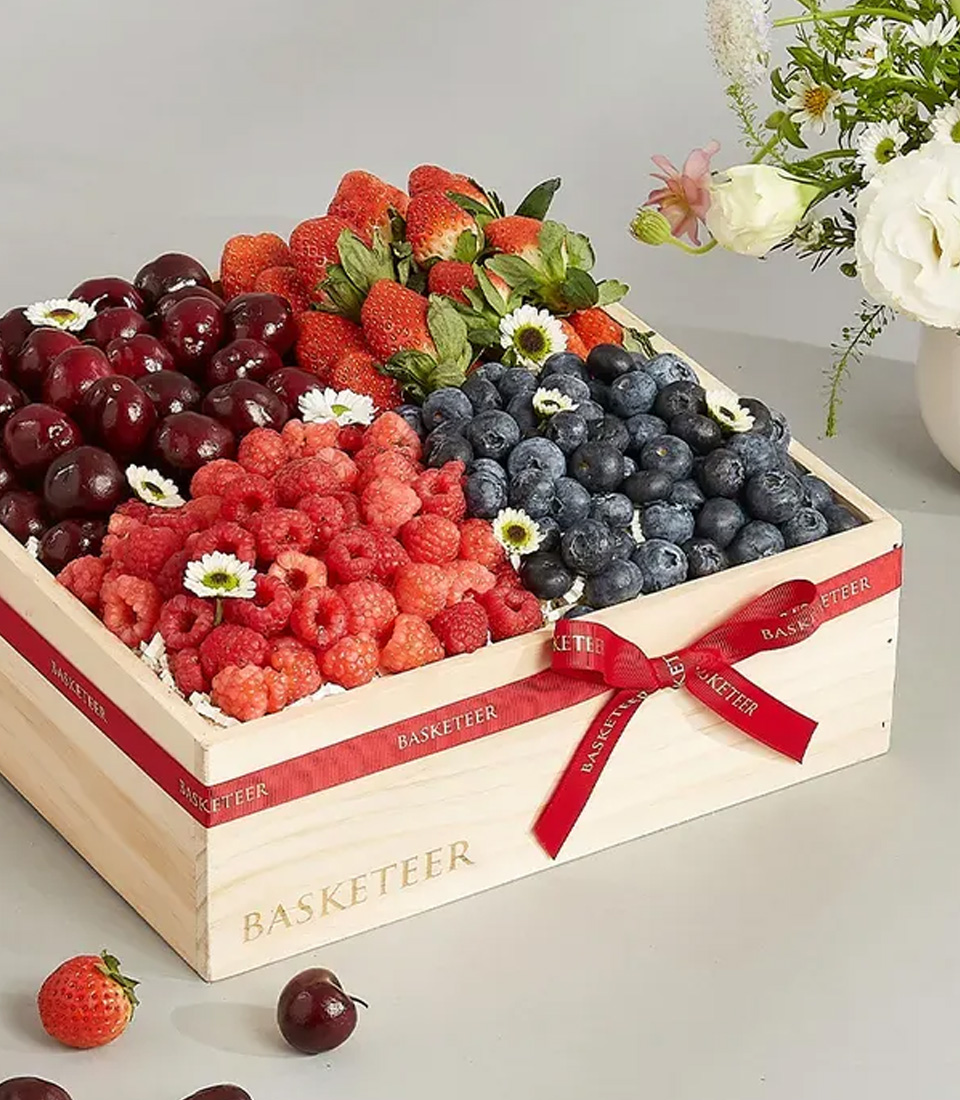 A wooden gift crate filled with fresh cherries, strawberries, raspberries, and blueberries, tied with a red ribbon, with a white flower vase in the background.