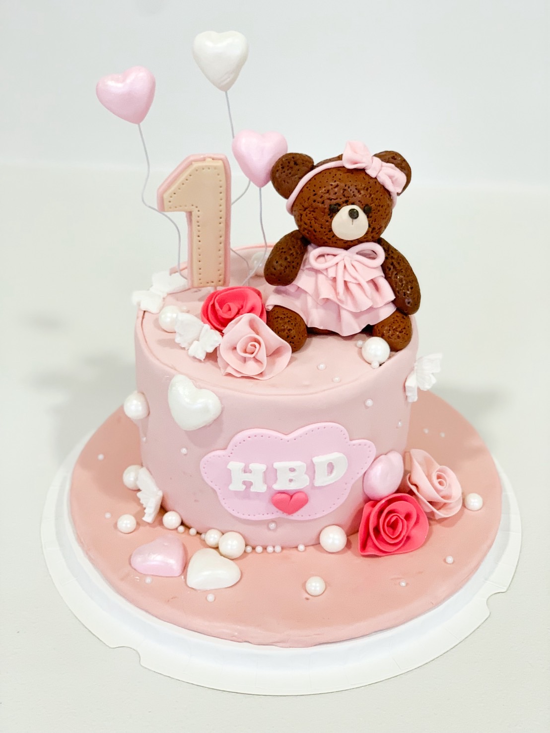 Pink first birthday cake decorated with fondant teddy bear, roses, pearls, hearts, and number 1 topper.