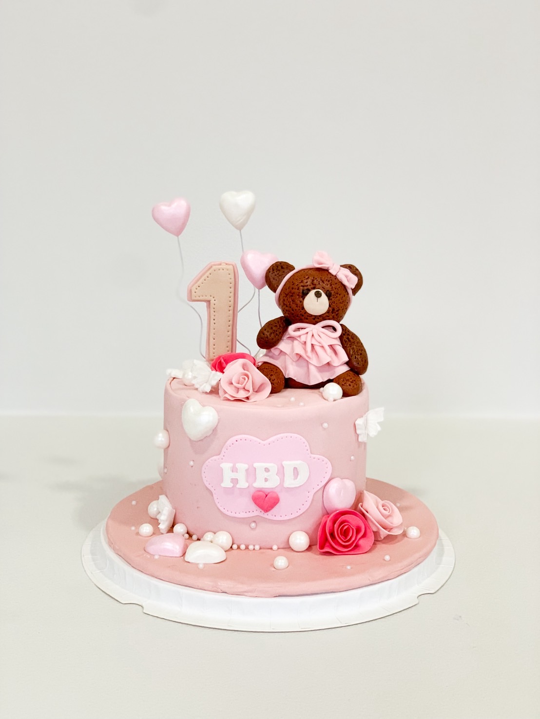 Pink first birthday cake with fondant teddy bear, roses, pearls, hearts, and number 1 topper.