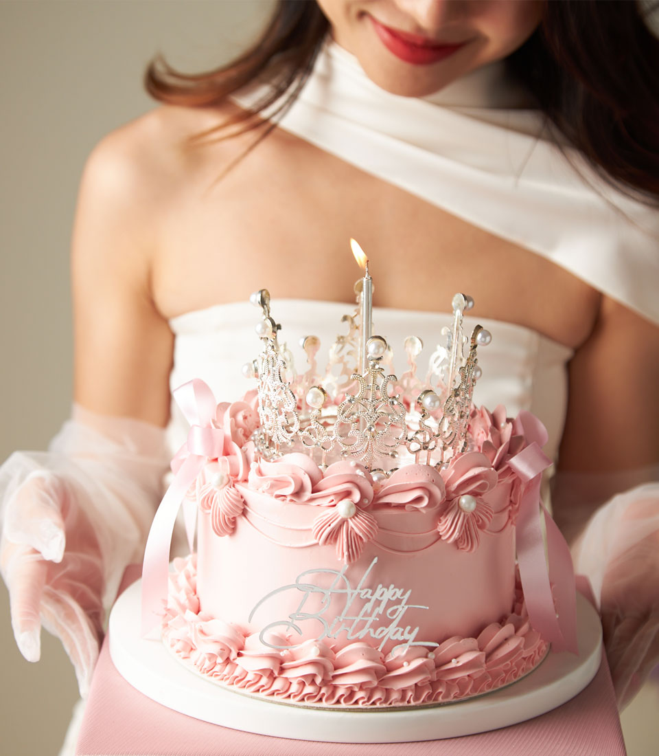 A woman in a white dress holds a pink birthday cake decorated with piped flowers, ribbons, a crown-shaped topper, and a single lit candle. The cake has "Happy Birthday" written on it.