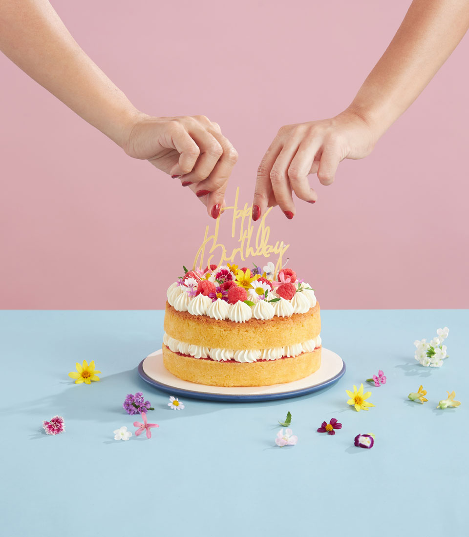 Two hands decorate a layered birthday cake with piped cream and colorful flowers on a blue table, with scattered flowers around. The background is pink.