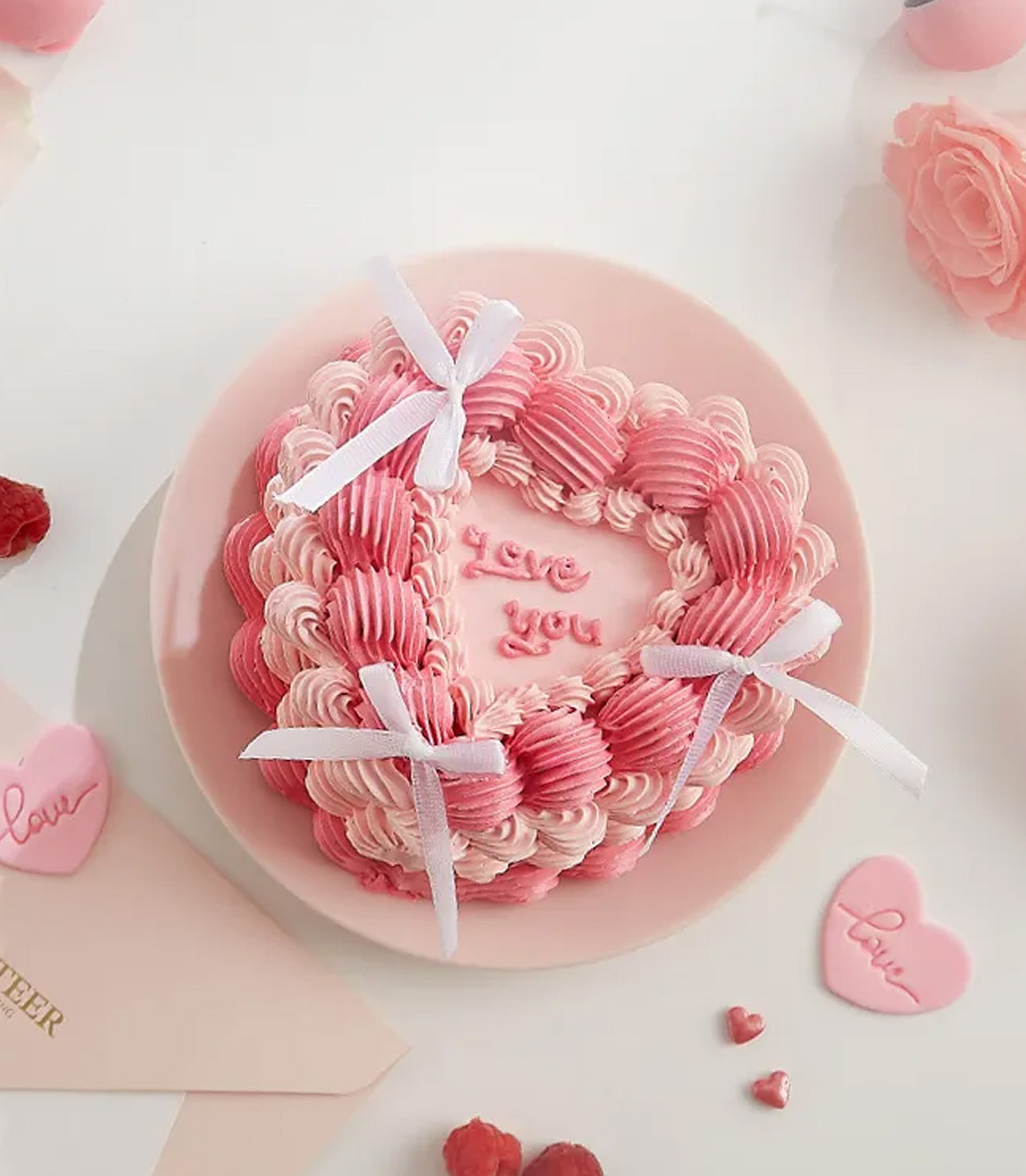 A heart-shaped pink and white cake with “Love you” written in the center, decorated with piped frosting, two white ribbons, and surrounded by roses and heart-shaped decorations on a pink plate.