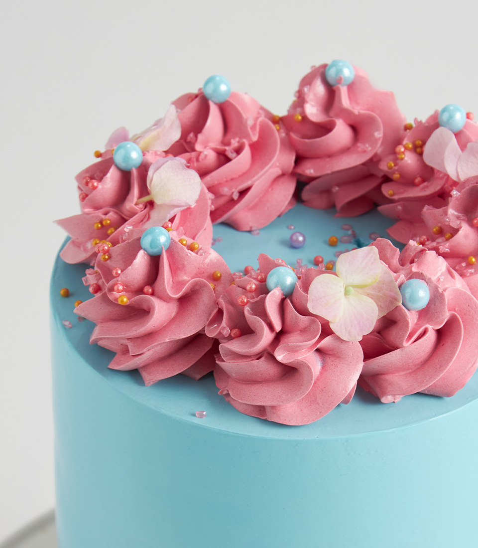Close-up of Blue cake topped with pink buttercream swirls, pastel pearls, and hydrangea petals