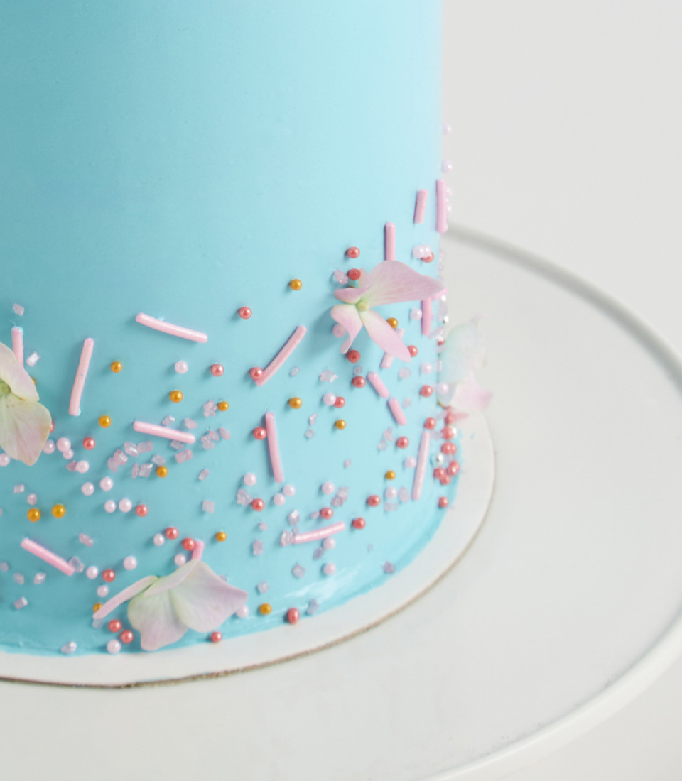 Close-up of Blue cake topped with pink buttercream swirls, pastel pearls, and hydrangea petals