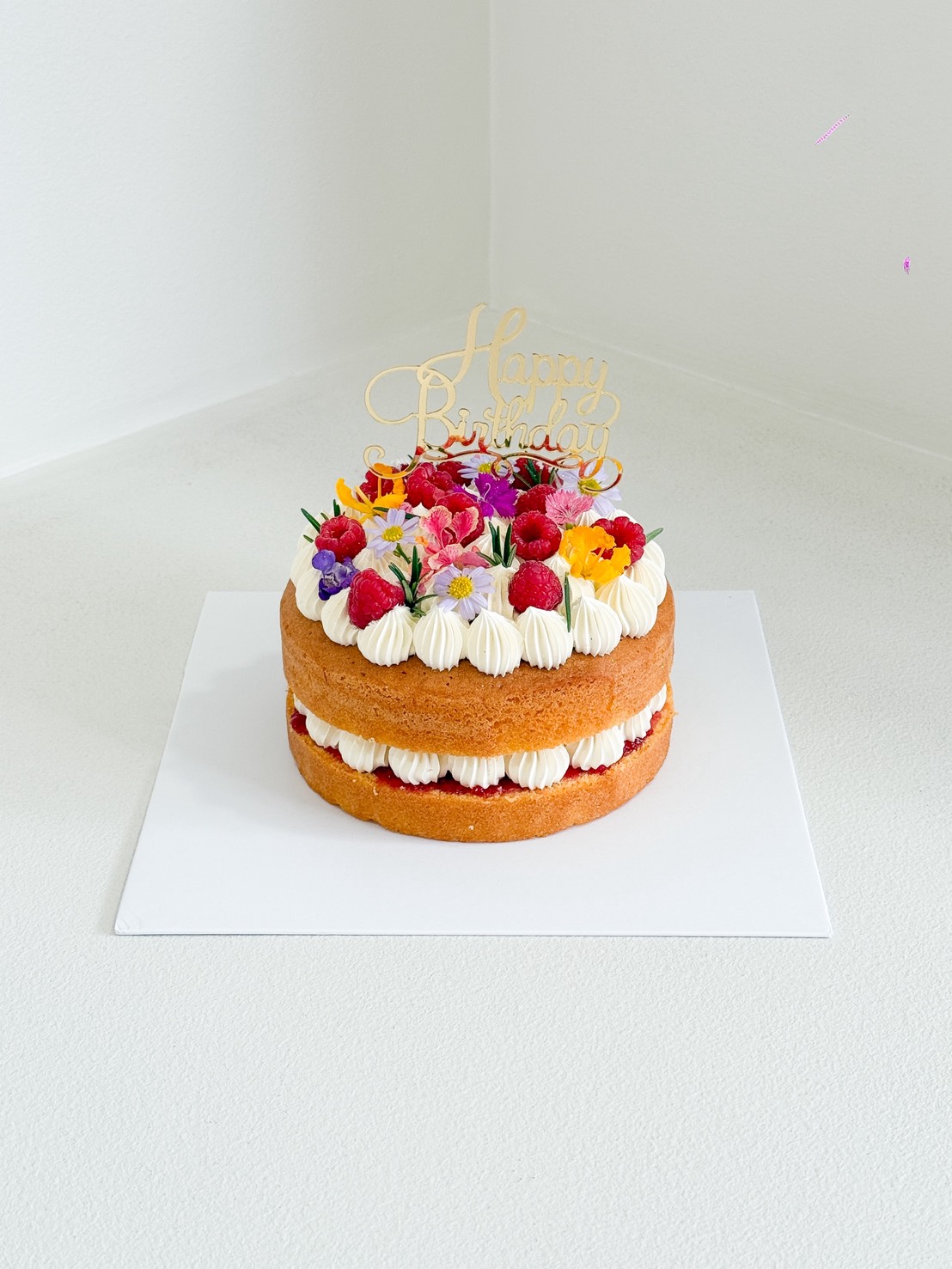A Raspberry Vanilla Naked Cake, this two-layer sponge cake with white icing and colourful edible flowers is displayed on a white board. A gold 