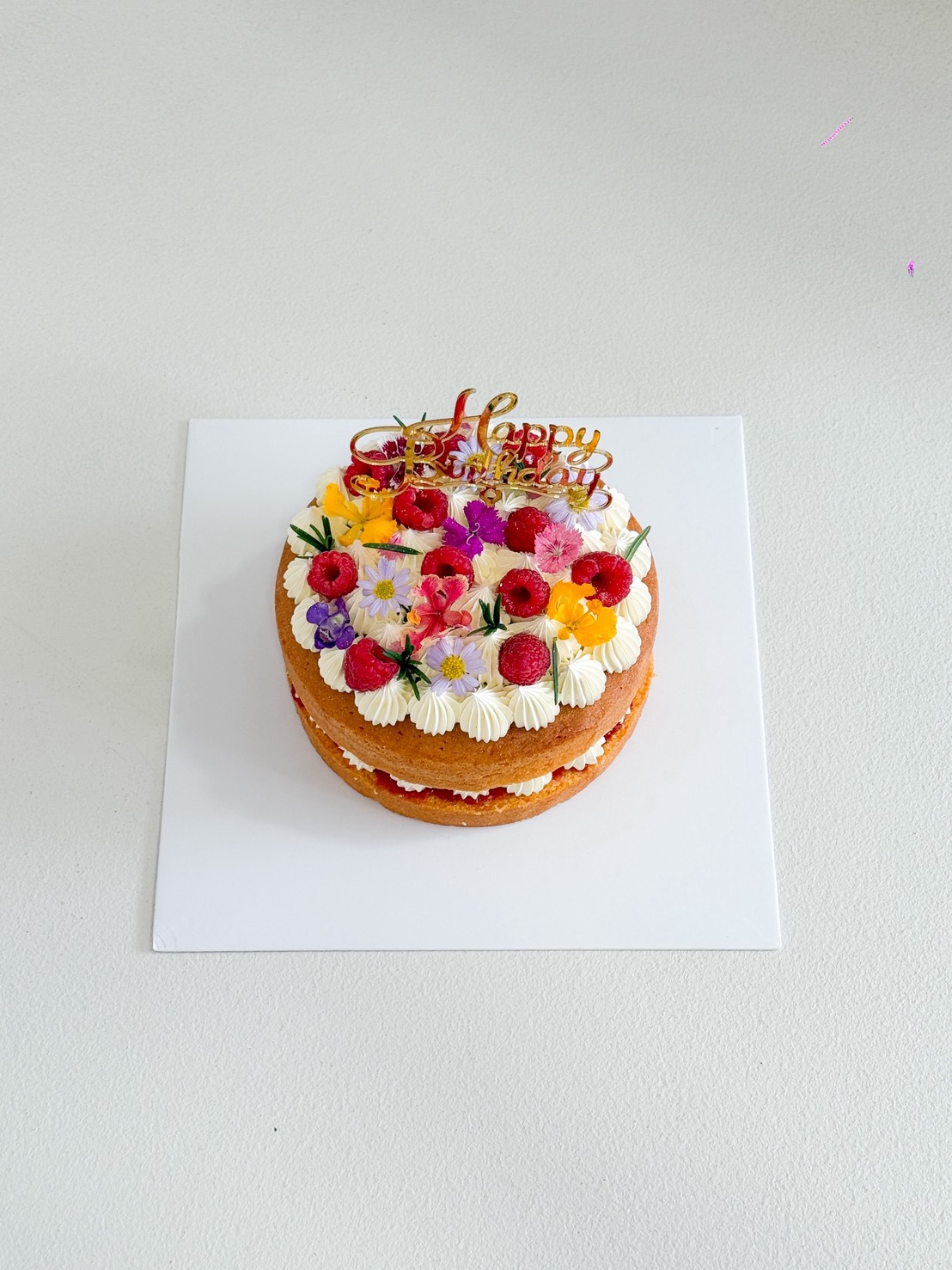 A Raspberry Vanilla Naked Cake topped with white icing, fresh raspberries, and colourful edible flowers sits on a white square board with a gold 