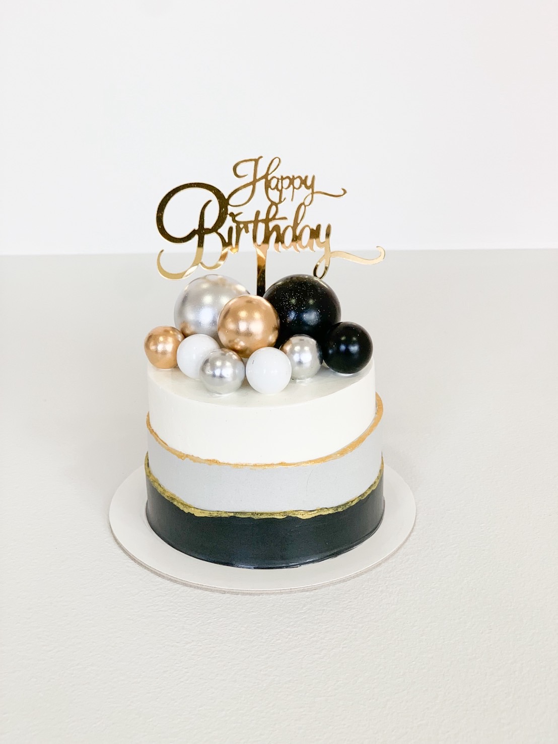 Round birthday cake with black, grey, and white frosting, topped with gold, silver, and black edible spheres and a 