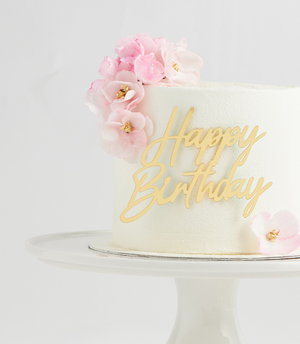 Elegant white cake with pink flowers and gold “Happy Birthday” topper on a pedestal stand