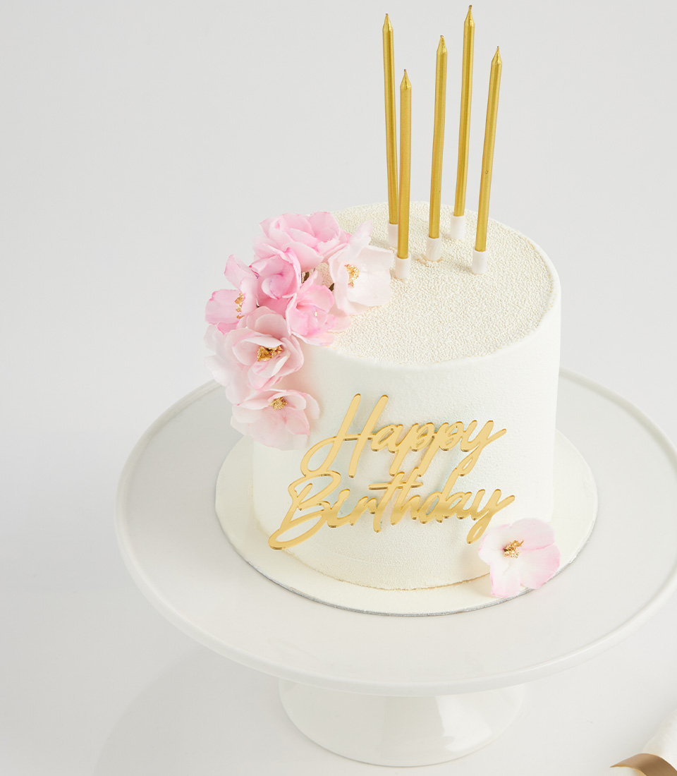 White birthday cake topped with pink wafer flowers and gold candles