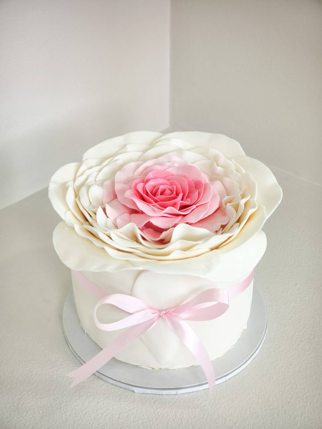 Elegant fondant cake topped with large handcrafted rose petals in white and pink tones, tied with a soft pink ribbon