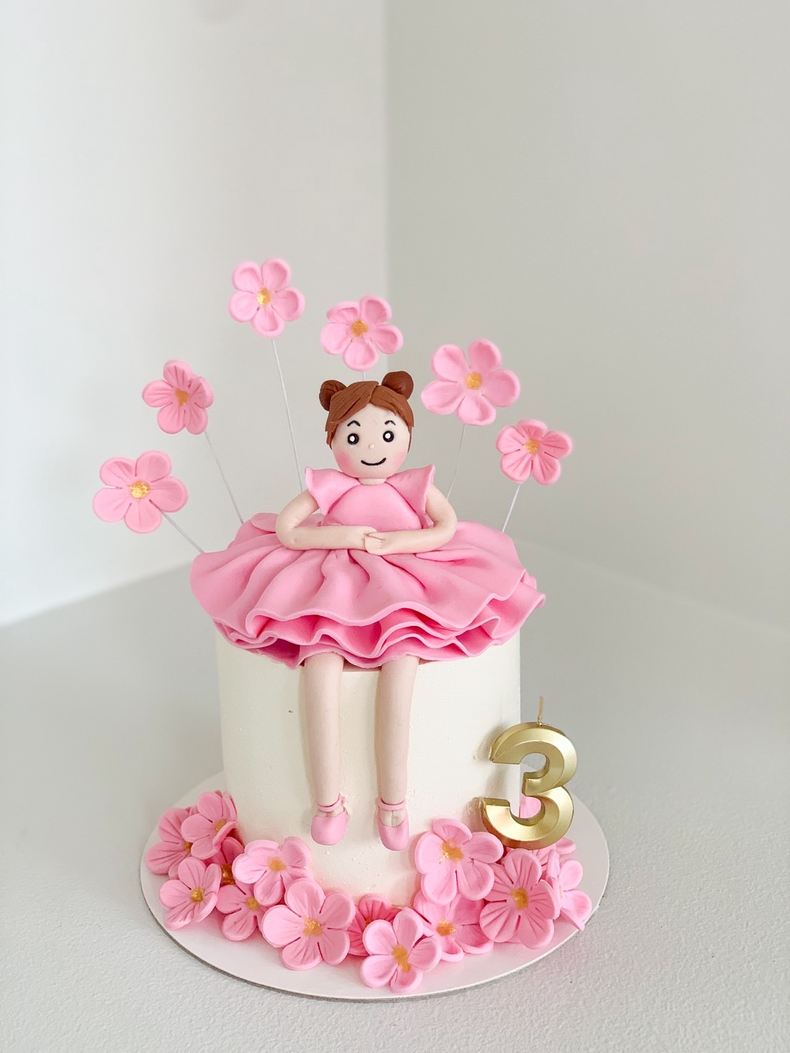 White round birthday cake decorated with a pink ballerina girl topper, pink flowers, and a gold number 3 candle.