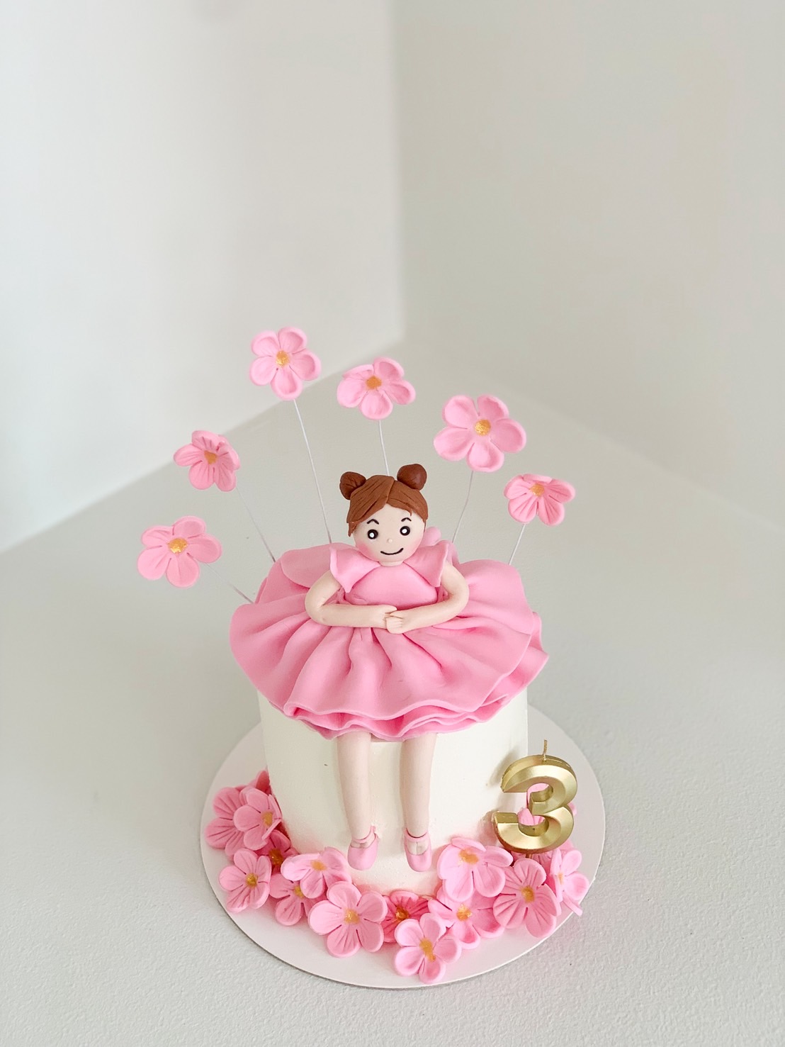 White round birthday cake decorated with a pink ballerina girl topper, pink flowers, and a gold number 3 candle.
